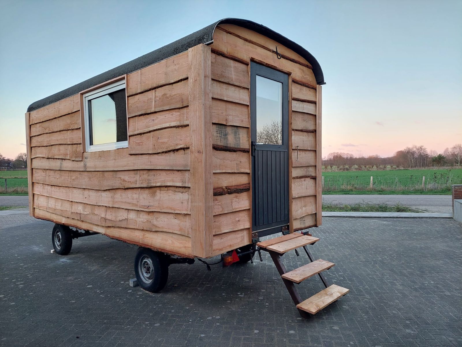 Pipowagen met ruw gezaagde houten gevelbekleding, gebogen dak, zwarte deur en houten trapje
