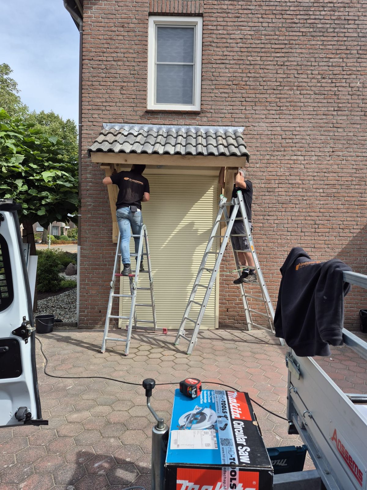 Twee mannen op ladders plaatsen een op maat gemaakt dakje met dakpannen boven een voordeur